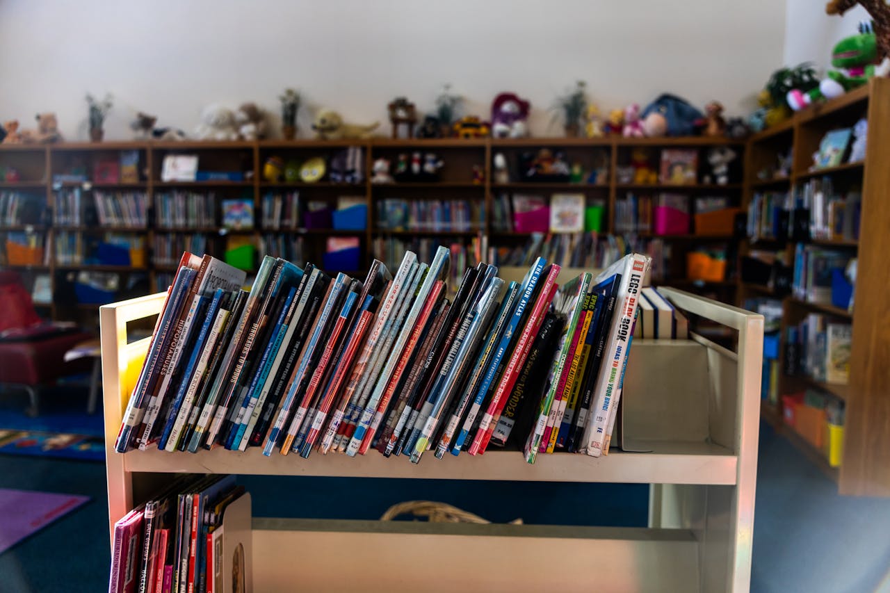 A warm library interior featuring a book cart in focus with colorful children's books on shelves.